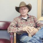 Joe Merrick, the local cowboy and musical artist, seated in front of a grey wall wearing a cowboy hat, long sleeve shirt, and jeans.